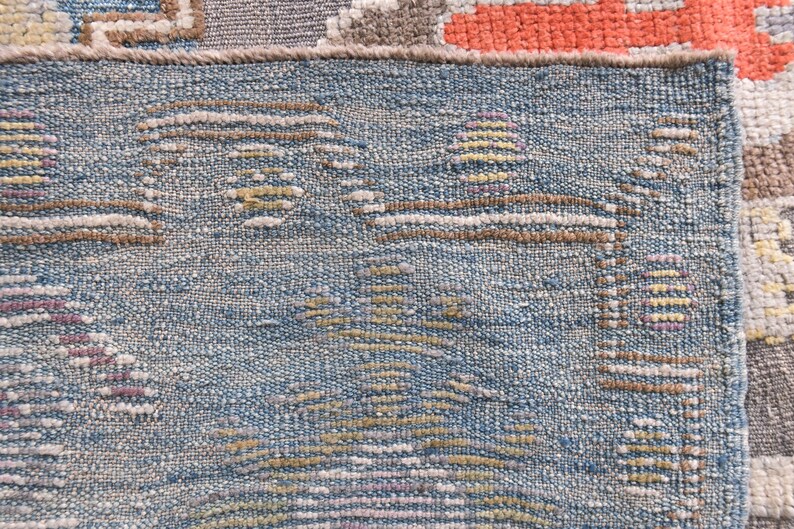 May include: Close-up of a section of a woven rug with a blue background and geometric patterns in brown, yellow, and purple. The rug has a textured appearance, with visible threads and a detailed design. The rug's edge is visible.
