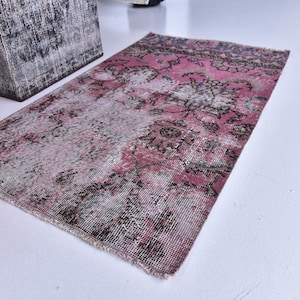 Könnte beinhalten: Ein rechteckiger, verwaschener rosa Teppich mit floralem Muster. Der Teppich hat ein verblasstes Aussehen mit Rosa-, Grau- und Schwarztönen. Der Teppich liegt auf einer weißen Oberfläche.