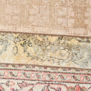 May include: Close-up of the bottom edge of a rug with a beige background and a patterned border. The border features a repeating design of pink, green, and brown.