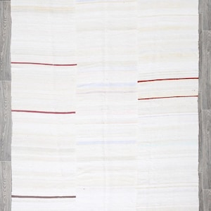 May include: An off-white rectangular rug with a patchwork design. The rug has horizontal stripes in shades of white and beige, with thin, dark red and brown accent lines. The rug is on a grey wooden floor.