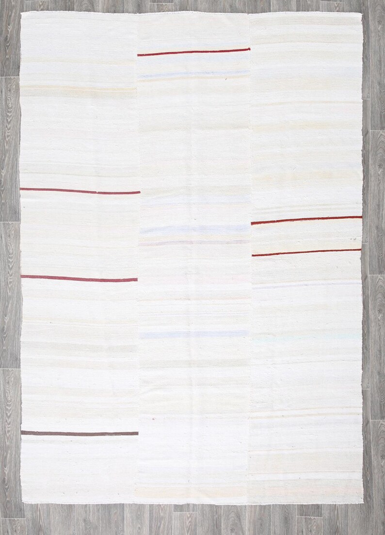 May include: An off-white rectangular rug with a patchwork design. The rug has horizontal stripes in shades of white and beige, with thin, dark red and brown accent lines. The rug is on a grey wooden floor.