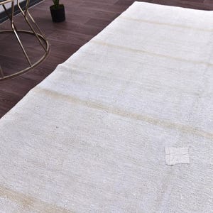 Peut inclure: Un grand tapis rectangulaire blanc cassé avec des rayures beiges horizontales. Le tapis a une surface texturée et quelques petits patchs. Le tapis est posé sur un sol en bois brun foncé, avec une table d'appoint en métal doré en arrière-plan.
