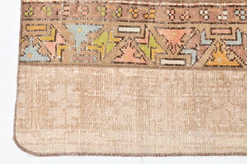 May include: Close-up of the bottom edge of a vintage rug with a brown fringe. The rug has a light brown background with a colorful geometric pattern.