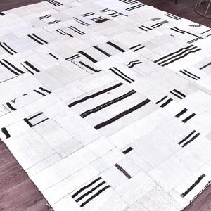 May include: A rectangular, patchwork rug with a white background and black geometric designs. The rug features various rectangular and square patches, each with black stripes or solid black shapes. The rug is placed on a wooden floor.