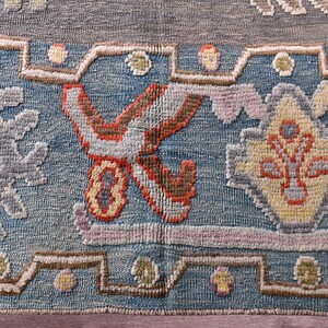 May include: Close-up of a section of a rug with a detailed, geometric pattern. The rug features a blue background with various shapes in shades of grey, pink, yellow, and orange. The design includes floral and geometric motifs.
