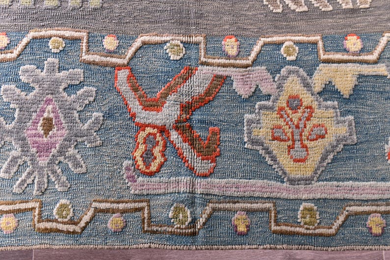 May include: Close-up of a section of a rug with a detailed, geometric pattern. The rug features a blue background with various shapes in shades of grey, pink, yellow, and orange. The design includes floral and geometric motifs.