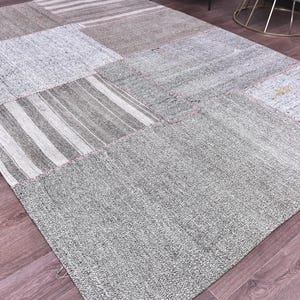 May include: A patchwork rug with a variety of gray and beige tones. The rug is made up of rectangular and square patches with different textures and patterns, including stripes. The rug is placed on a wooden floor.
