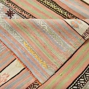 May include: A close-up of a woven rug with a striped pattern in shades of pink, green, blue, and white. The rug has a fringe border and intricate geometric designs.
