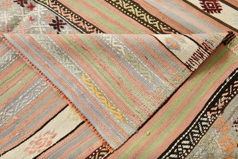 May include: A close-up of a woven rug with a striped pattern in shades of pink, green, blue, and white. The rug has a fringe border and intricate geometric designs.