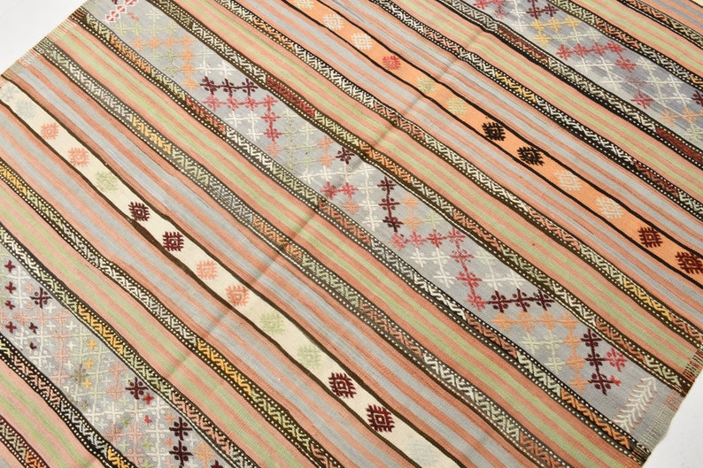 May include: A traditional Turkish kilim rug with a striped pattern in shades of pink, beige, brown, and gray. The rug features intricate geometric designs woven into the fabric.
