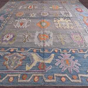 May include: A large, rectangular rug with a blue and grey background. It features a repeating pattern of floral and geometric designs in orange, purple, and yellow. The border is a darker blue, with a patterned design.