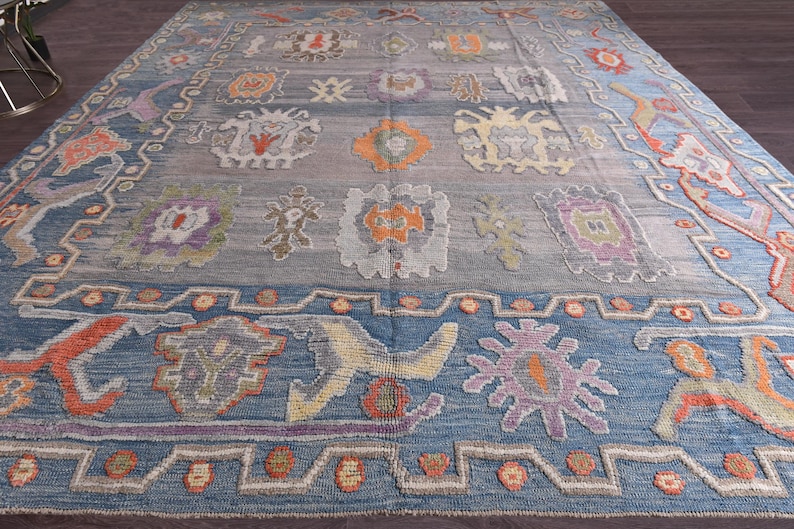 May include: A large, rectangular rug with a blue and grey background. It features a repeating pattern of floral and geometric designs in orange, purple, and yellow. The border is a darker blue, with a patterned design.