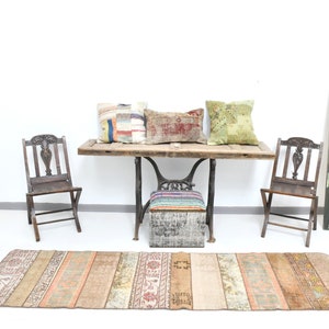 May include: A rustic wooden table with a metal base, featuring three colorful throw pillows and two wooden chairs. A vintage rug with a striped pattern is placed in front of the table. A painting of a woman in a yellow and green striped dress hangs on the wall.