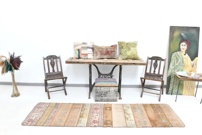 May include: A rustic wooden table with a metal base, featuring three colorful throw pillows and two wooden chairs. A vintage rug with a striped pattern is placed in front of the table. A painting of a woman in a yellow and green striped dress hangs on the wall.