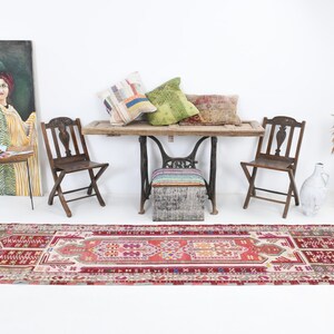 K&ouml;nnte beinhalten: Ein Vintage-L&auml;ufer mit einem geometrischen Muster in Rot, Rosa und Wei&szlig;. Der L&auml;ufer liegt auf einem wei&szlig;en Boden in einem Raum mit einem Holztisch, zwei St&uuml;hlen und einem Gem&auml;lde an der Wand.