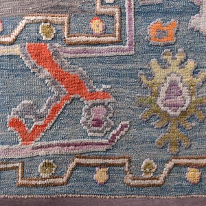 May include: Detail of a blue rug with a complex pattern. The rug incorporates orange, yellow, purple, and grey, forming a detailed, textured design. The rug's design has a floral theme. The rug is likely made of wool or a similar material.