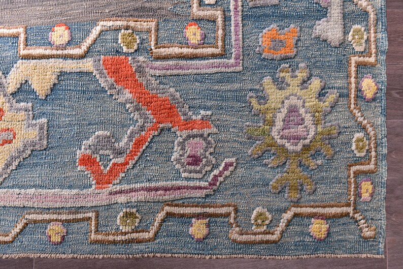 May include: Detail of a blue rug with a complex pattern. The rug incorporates orange, yellow, purple, and grey, forming a detailed, textured design. The rug's design has a floral theme. The rug is likely made of wool or a similar material.