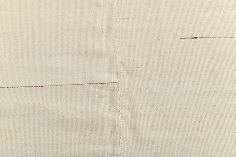 May include: Close-up of a plain, off-white fabric with a subtle texture. The fabric has two horizontal seams running across it.