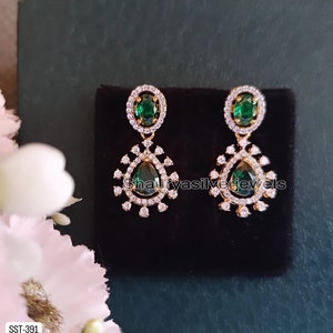 Pu&ograve; includere: Un paio di orecchini color oro con gemme a goccia verde smeraldo. Ogni orecchino presenta una gemma verde ovale pi&ugrave; piccola nella parte superiore, circondata da piccole pietre bianche. Gli orecchini sono esposti su una superficie nera.