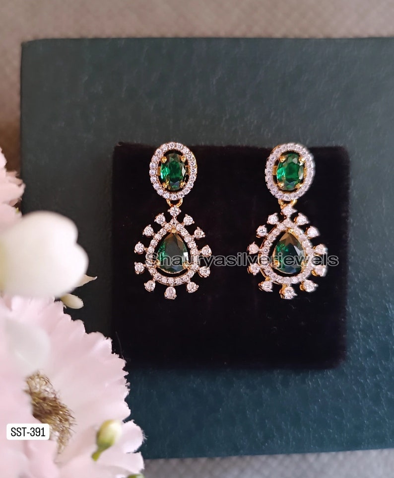 Pu&ograve; includere: Un paio di orecchini color oro con gemme a goccia verde smeraldo. Ogni orecchino presenta una gemma verde ovale pi&ugrave; piccola nella parte superiore, circondata da piccole pietre bianche. Gli orecchini sono esposti su una superficie nera.