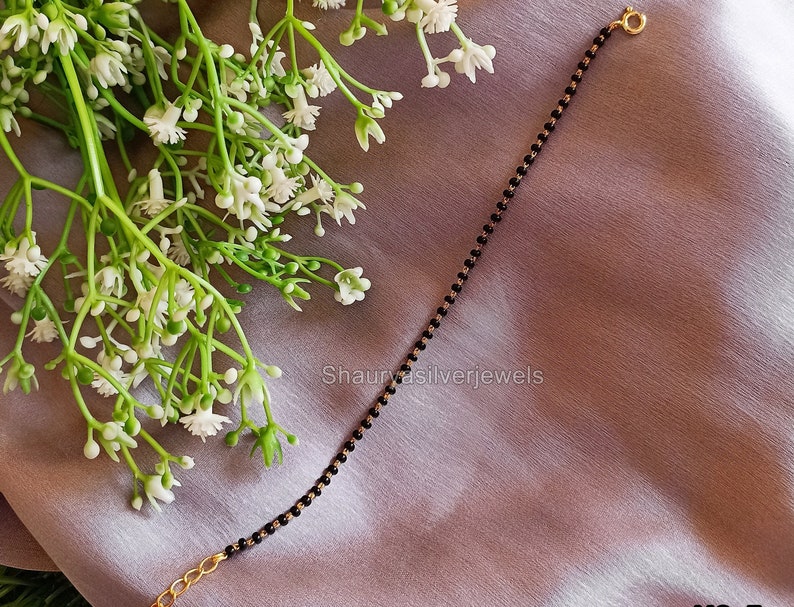 Peut inclure: Un bracelet d&eacute;licat &agrave; perles noires avec des maillons et un fermoir dor&eacute;s. Le bracelet est pr&eacute;sent&eacute; sur un tissu doux, violet clair, avec des fleurs blanches et des tiges vertes en arri&egrave;re-plan. Le texte "Shaurya silverjewels" est visible.