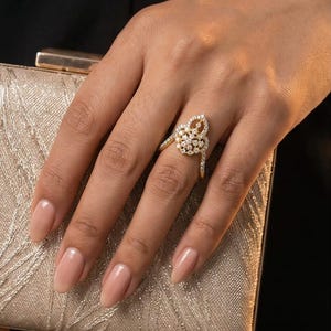 Op de afbeelding: Een gouden ring met een druppelvormige cluster van kleine heldere steentjes in een bloemmotief, met extra steentjes ingelegd langs de ringband. De ring wordt getoond tegen een gestructureerde gouden clutch met de tekst SST-596 zichtbaar.
