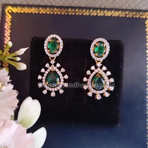 Pu&ograve; includere: Un paio di orecchini color oro con pietre preziose verdi smeraldo a forma di goccia. Ogni orecchino presenta un alone di piccole pietre trasparenti attorno alla pietra verde centrale e un design pendente decorativo. Gli orecchini sono esposti su una superficie nera.
