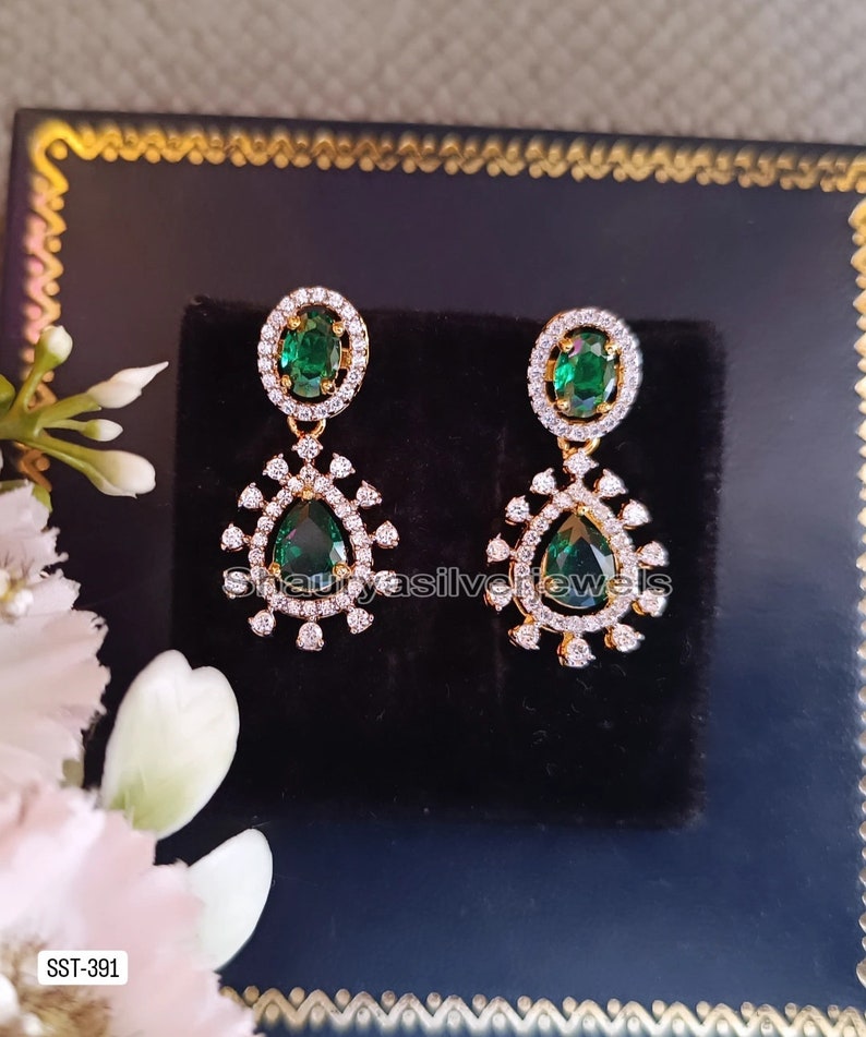 Pu&ograve; includere: Un paio di orecchini color oro con pietre preziose verdi smeraldo a forma di goccia. Ogni orecchino presenta un alone di piccole pietre trasparenti attorno alla pietra verde centrale e un design pendente decorativo. Gli orecchini sono esposti su una superficie nera.