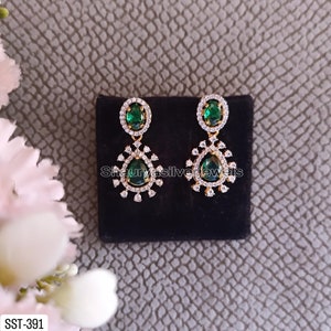 Pu&ograve; includere: Un paio di orecchini pendenti color oro con pietre preziose ovali e a goccia verde smeraldo. Gli orecchini sono impreziositi da piccoli cristalli trasparenti e sono esposti su una superficie nera. Il testo "Shaurya silver jewels" &egrave; visibile.