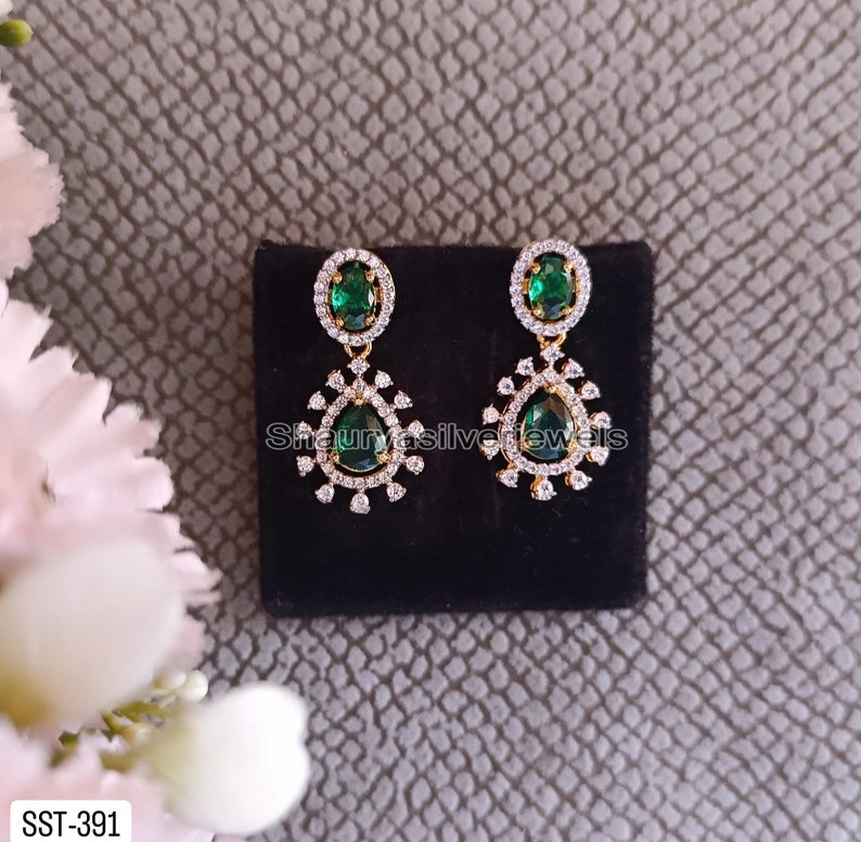 Pu&ograve; includere: Un paio di orecchini pendenti color oro con pietre preziose ovali e a goccia verde smeraldo. Gli orecchini sono impreziositi da piccoli cristalli trasparenti e sono esposti su una superficie nera. Il testo "Shaurya silver jewels" &egrave; visibile.