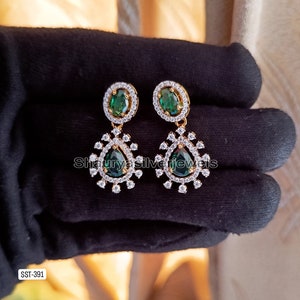 Pu&ograve; includere: Orecchini pendenti color oro con pietre ovali e a goccia verde smeraldo. Gli orecchini sono impreziositi da piccoli cristalli trasparenti. Il testo "Shauryasilverjewels" &egrave; visibile. Gli orecchini sono esposti su una superficie nera.