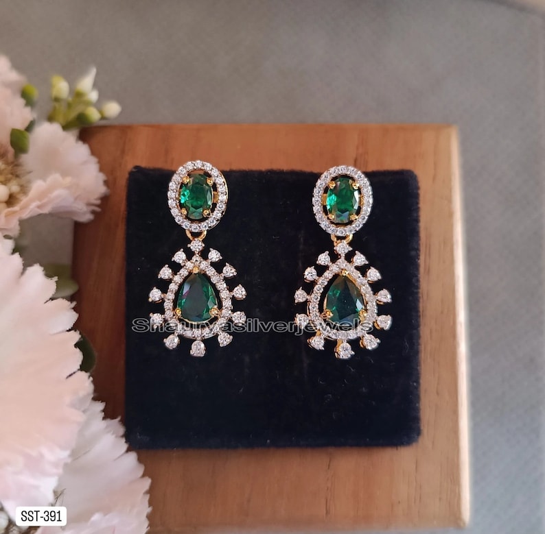 Pu&ograve; includere: Un paio di orecchini pendenti color oro con accenti di smeraldo verde e cristalli trasparenti. Ogni orecchino presenta una pietra verde ovale circondata da cristalli trasparenti nella parte superiore e una pietra verde a forma di pera nella parte inferiore.