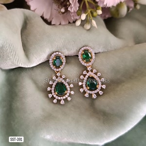 Pu&ograve; includere: Orecchini pendenti color oro con pietre centrali a goccia verde smeraldo e pietre ovali pi&ugrave; piccole. Gli orecchini sono circondati da un alone di piccole pietre trasparenti. Esposti su una superficie verde chiaro.