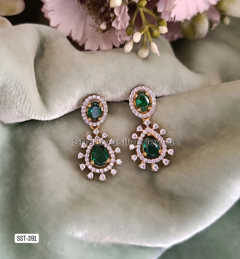 Pu&ograve; includere: Orecchini pendenti color oro con pietre centrali a goccia verde smeraldo e pietre ovali pi&ugrave; piccole. Gli orecchini sono circondati da un alone di piccole pietre trasparenti. Esposti su una superficie verde chiaro.