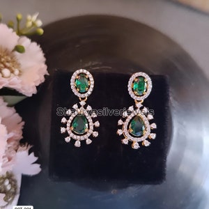 Pu&ograve; includere: Un paio di orecchini pendenti color oro con dettagli in verde smeraldo e cristallo trasparente. Gli orecchini hanno una pietra superiore ovale e una pietra inferiore a goccia. Sono esposti su una superficie nera.