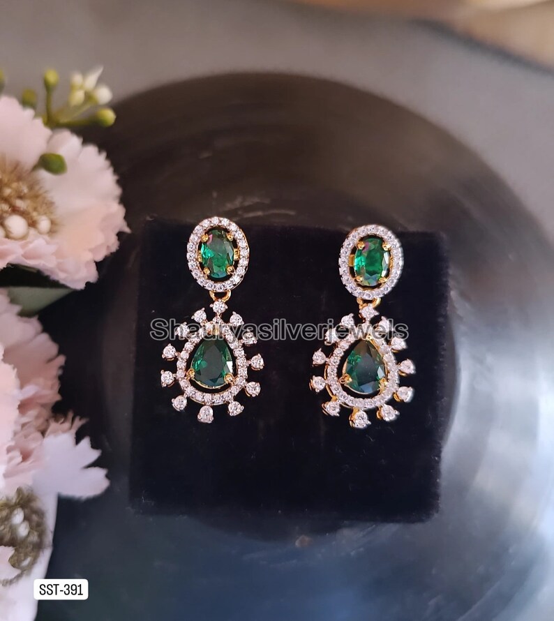 Pu&ograve; includere: Un paio di orecchini pendenti color oro con dettagli in verde smeraldo e cristallo trasparente. Gli orecchini hanno una pietra superiore ovale e una pietra inferiore a goccia. Sono esposti su una superficie nera.