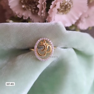 May include: Gold ring with a clear stone encrusted circle featuring an Om symbol. The ring is on a light green velvet surface. The text "SST-401" is in the bottom left corner of the image.