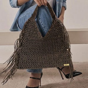 May include: A dark brown crochet tote bag featuring a V-shaped top opening, long fringe detailing along both side edges, and a small rectangular gold-toned metal brand tag attached to the lower right corner with the text Maffia.