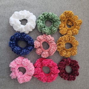 May include: Nine plush scrunchies in various colors, including white, green, gold, navy, pink, and burgundy. These soft, textured hair accessories are arranged on a gray surface, showcasing their vibrant hues and plush design.