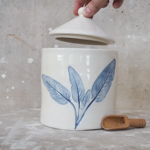 May include: A white ceramic canister with a lid, featuring a blue botanical leaf design. A small wooden scoop rests beside the canister. The canister is cylindrical and suitable for kitchen storage.