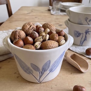 Könnte beinhalten: Eine weiße Keramikschale mit zwei kleinen Griffen und einem blauen Salbeiblatt-Design, gefüllt mit einer Auswahl an ganzen Nüssen wie Walnüssen, Haselnüssen, Mandeln und Pistazien. Die Schale hat eine glatte Oberfläche und runde Form.
