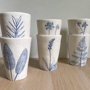 Op de afbeelding: Zes witte keramische bekers met blauwe botanische ontwerpen. De bekers hebben verschillende plantenillustraties, waaronder bladeren en bloemen. De bekers zijn gestapeld op een houten oppervlak.