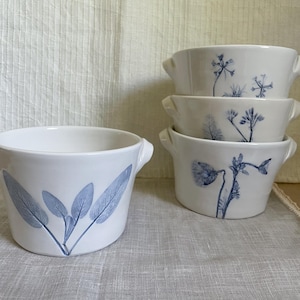 Op de afbeelding: Witte keramische kommen met blauwe botanische ontwerpen. Eén kom heeft saliebladeren, terwijl de andere verschillende bloempatronen tonen. Elke kom heeft twee kleine handvatten. De kommen zijn gerangschikt op een neutraal oppervlak.