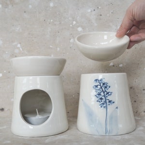 Könnte beinhalten: Weißer Keramik-Ölbrenner mit abnehmbarem Schälchen und einem kleinen Loch für ein Teelicht. Der Brenner hat ein florales Design mit blauen Blumen und Blättern.