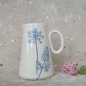 May include: A white ceramic pitcher with a blue floral design. The pitcher has a wide base and a handle. The floral design features a large flower and a smaller leaf.