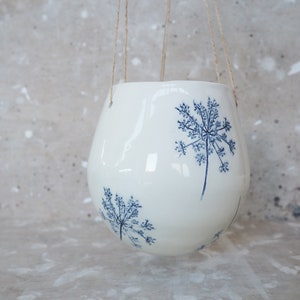 Può includere: Un vaso sospeso in ceramica bianca con finitura lucida. Il vaso presenta un design floreale blu ed è sospeso con spago naturale. La forma sferica e il motivo botanico creano un'estetica moderna e minimalista.