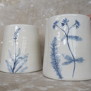 Op de afbeelding: Twee witte keramische vazen met een glanzende afwerking. Elke vaas heeft een handgeschilderd blauw botanisch ontwerp. Eén vaas heeft een lavendeltakje en de andere een varenachtige plant. De vazen hebben een licht uitlopende vorm.
