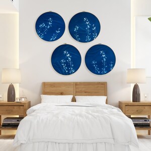 Op de afbeelding: Vier ronde wandkunstwerken met een diepblauwe achtergrond en witte bloemmotieven worden boven een bed getoond. De slaapkamer heeft een houten bedframe, twee nachtkastjes met lampen en een wit dekbed.