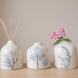 Può includere: Tre vasi in ceramica bianca con forme arrotondate e aperture strette. Ogni vaso presenta un'illustrazione botanica blu unica. Un vaso contiene bastoncini diffusori in legno, mentre un altro contiene dei piccoli fiori rosa.