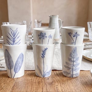 May include: A set of six white ceramic tumblers arranged in pairs, each featuring a unique blue botanical illustration. The designs include detailed depictions of leaves, lavender, and wildflowers, with a smooth finish and tapered cylindrical shape.